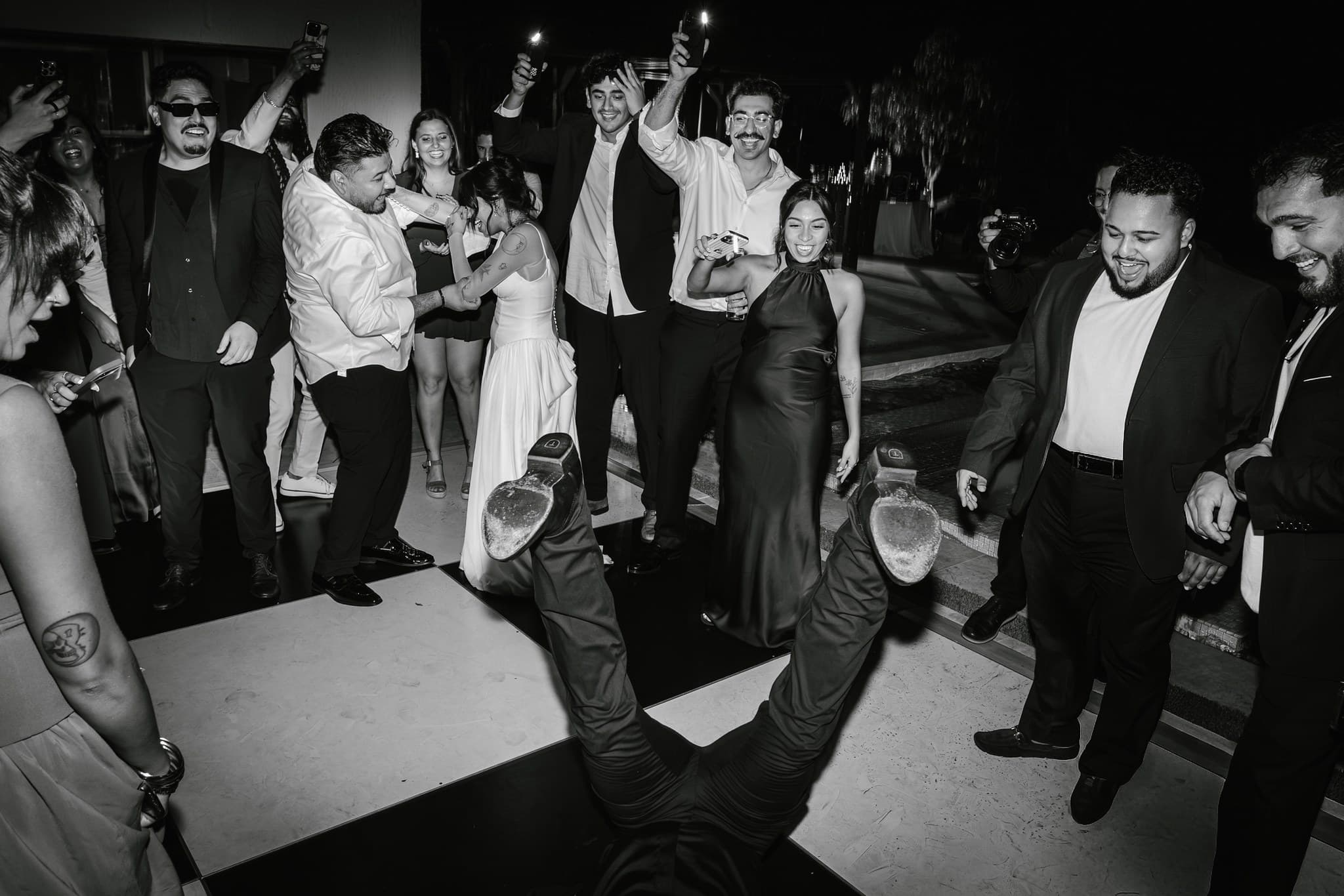Guests crowd a checkered dance floor as someone spins on their back, shoes in the air, while the wedding party cheers