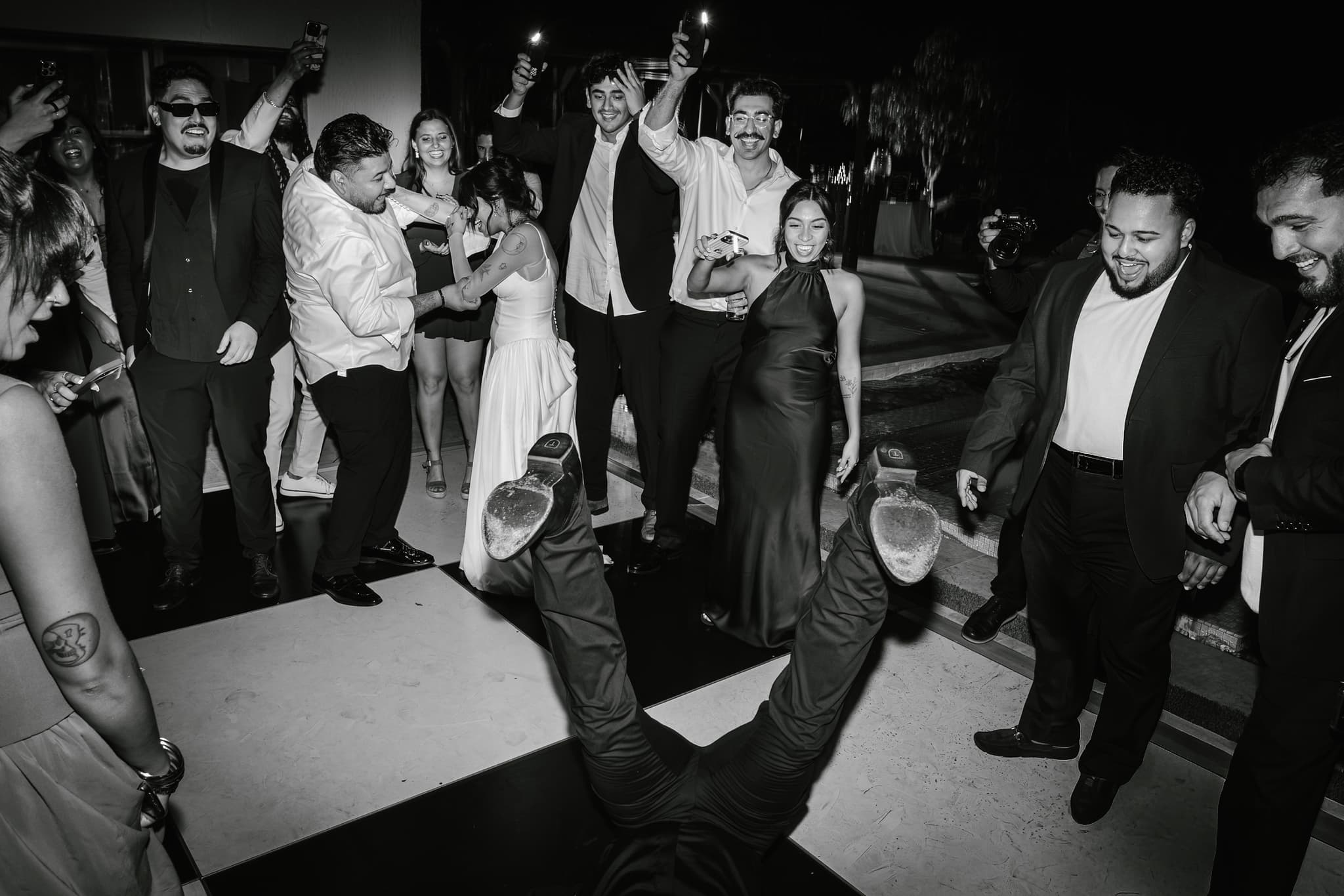 Guests crowd a checkered dance floor as someone spins on their back, shoes in the air, while the wedding party cheers