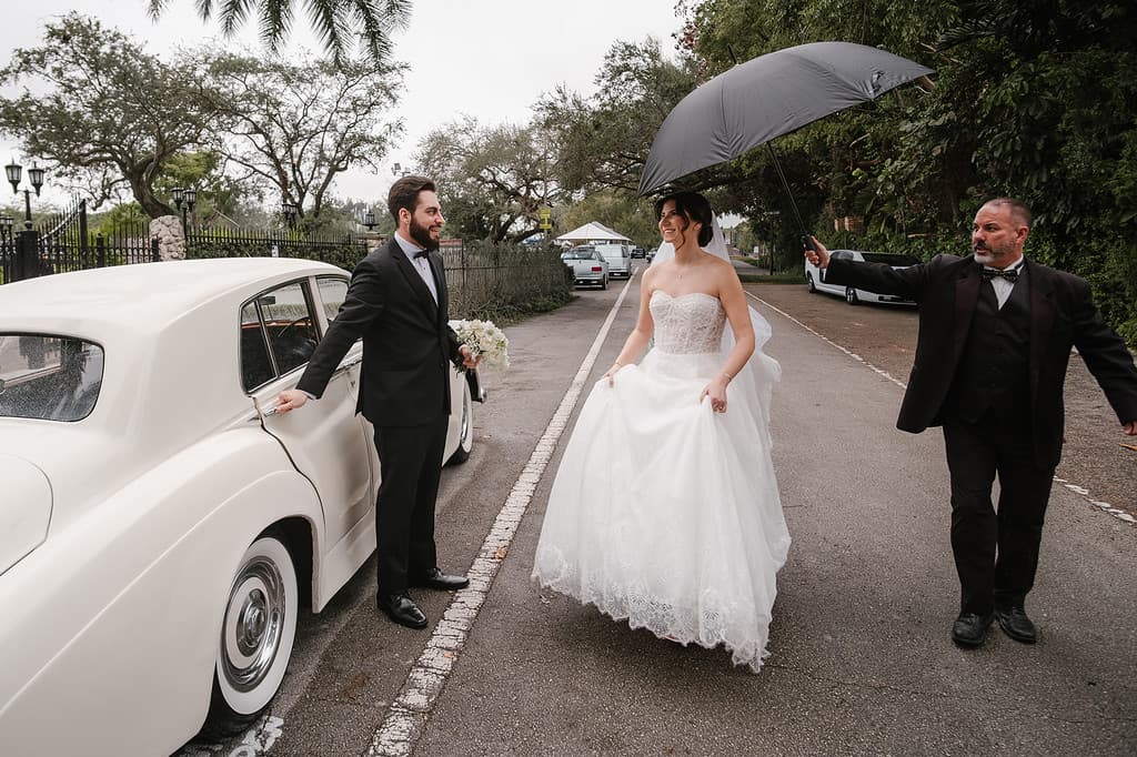 A bride and groom beside a light vintage car while guests stand nearby