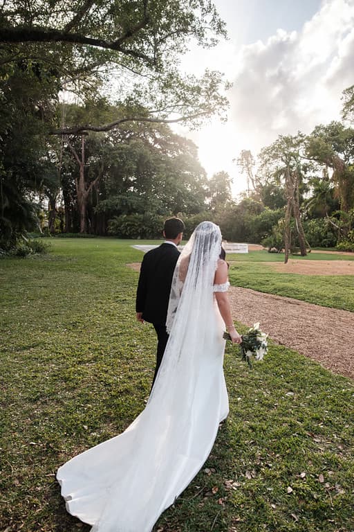 A bride and groom from behind on a lawn, her veil trailing in the light