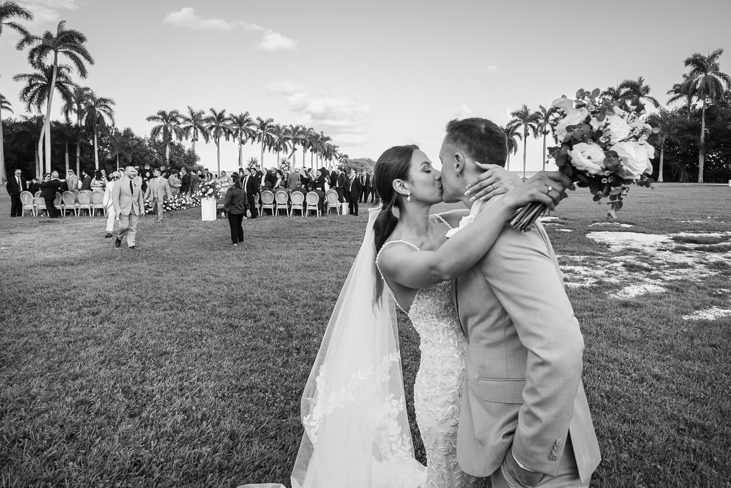A bride and groom embracing on a lawn with palm trees behind them