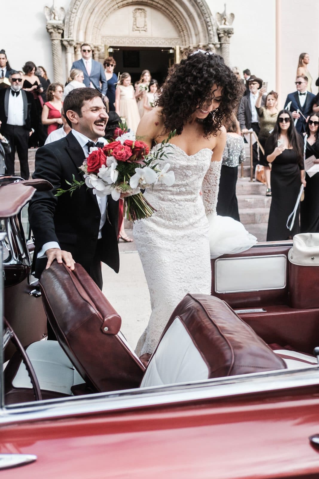 Sophia and Patrick beside a vintage car outside St. Patrick Catholic Church, Sophia holding a red and white bouquet