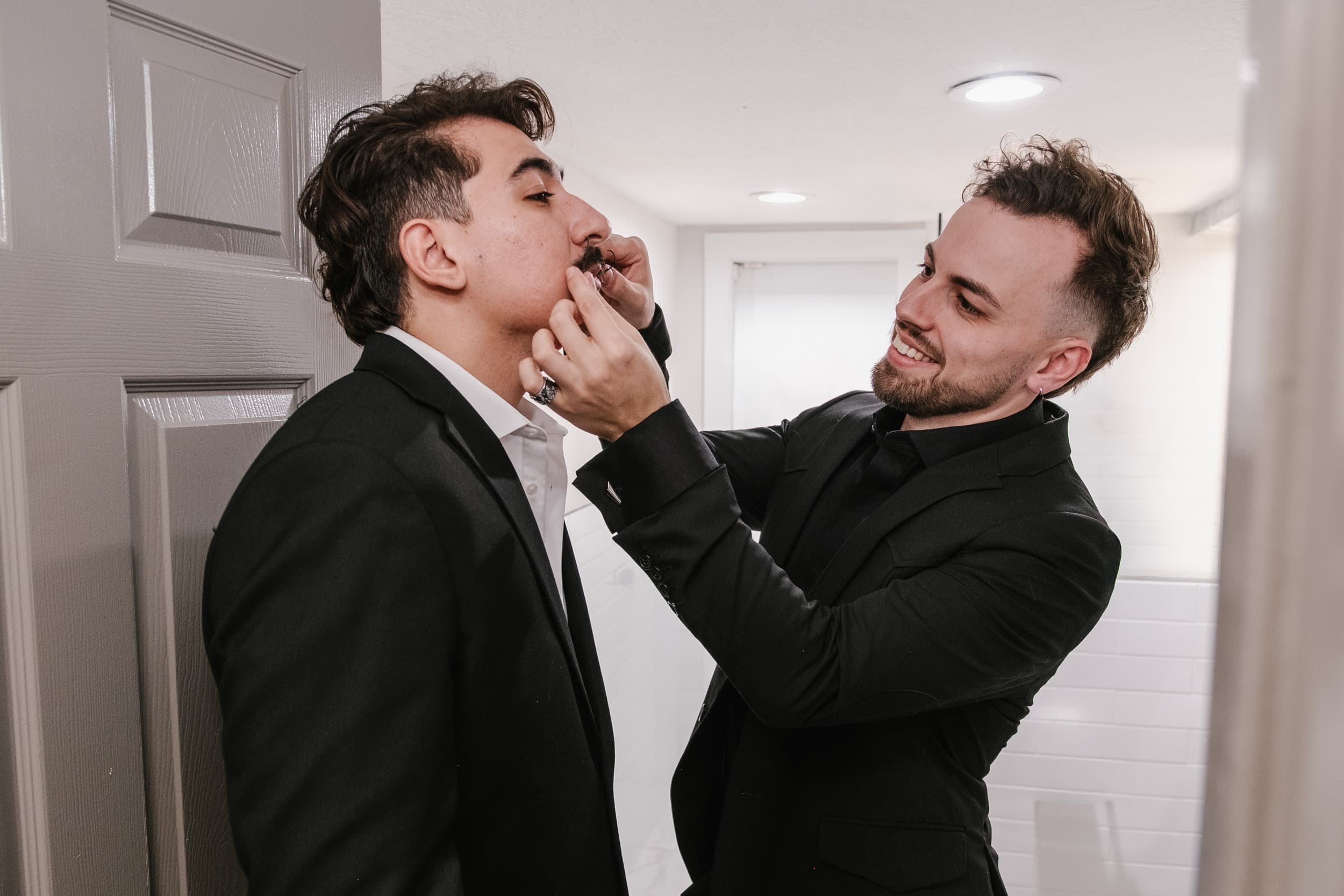 A groomsman laughs while fitting a mustache onto another man in a black suit in a hallway before the ceremony